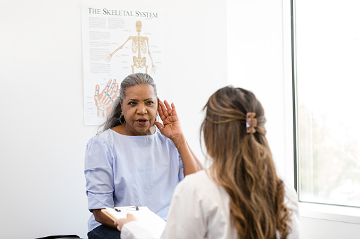 While the doctor is talking, the patient places her hand to her ear to let her know that she can't hear very well.