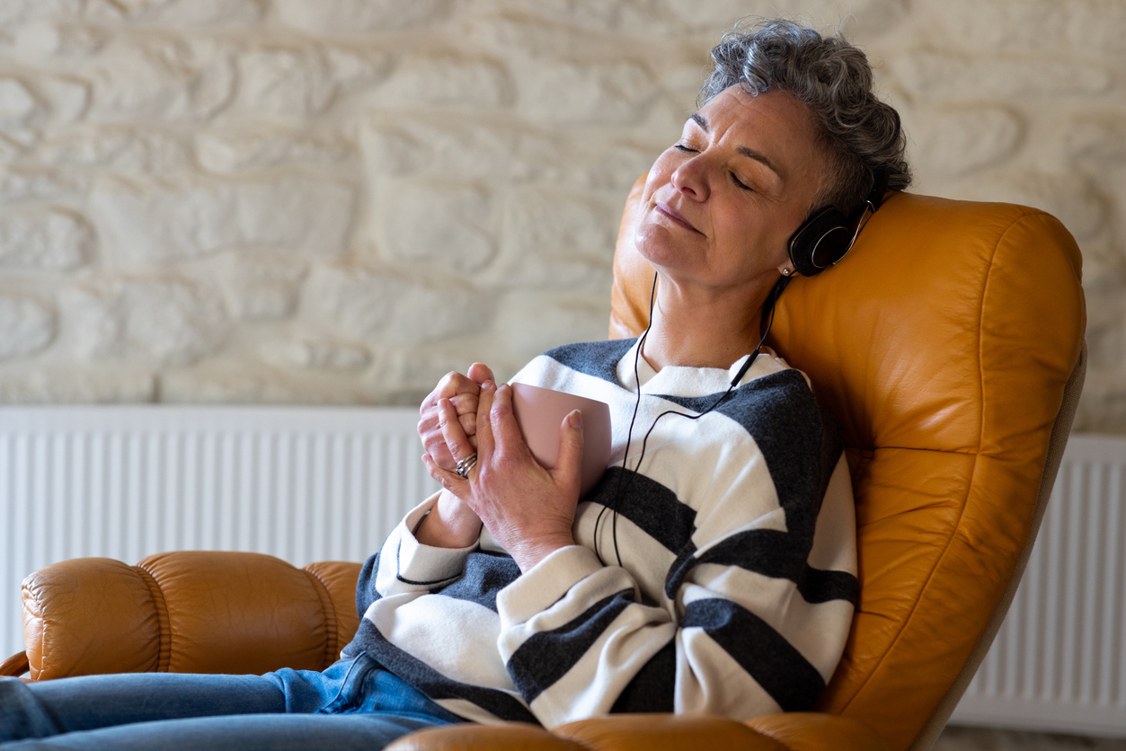Waist-up shot of a woman sitting in a lounge chair at home relaxing with her eyes closed. She is holding a hot drink and listening to music from overhead headphones.